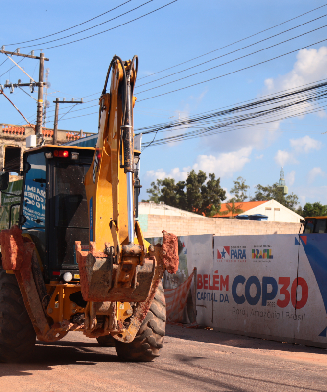 Uma retroescavadeira amarela realiza obras em uma rua de Belém, cercada por tapumes que exibem o logotipo do Governo do Pará e a frase “Belém, capital da COP 30 – Pará, Amazônia, Brasil”. O céu está azul e, ao fundo, há construções antigas e fios de energia cruzando a cena, indicando preparativos na cidade para receber o evento climático.