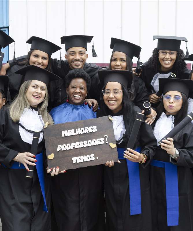 Um grupo diverso de formandos posa sorridente para a foto, usando becas e segurando diplomas. No centro, uma professora também sorri enquanto os abraça. Duas formandas à frente exibem uma placa com a frase: “Melhores professores? Temos.” O clima é de alegria, união e celebração pela conquista.