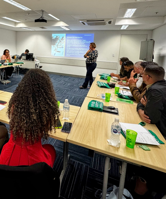 Participantes sentados em mesas organizadas em formato de “U” acompanham uma atividade formativa em uma sala de reunião iluminada. Uma facilitadora fala diante de uma apresentação projetada na parede, enquanto o grupo observa e faz anotações.