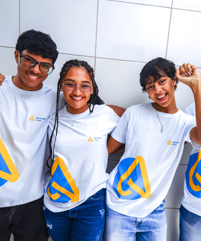 Quatro pessoas jovens estão em pé, lado a lado, com os braços entrelaçados pelos ombros, também em frente a uma parede clara de azulejos. Todas usam camisetas brancas do programa “Aprendiz Legal”, com o mesmo logotipo azul e amarelo.