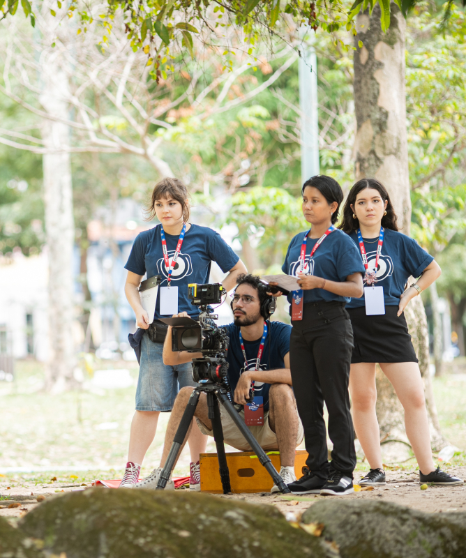 Um grupo de jovens realiza uma gravação ao ar livre, em um parque arborizado. À direita, uma equipe de audiovisual veste camisetas azuis e crachás de identificação; um dos integrantes está agachado atrás de uma câmera profissional montada em tripé, enquanto as colegas acompanham atentamente a cena. À esquerda, em primeiro plano, duas pessoas sentadas em um tronco conversam, uma delas segurando um violão, sugerindo uma entrevista ou apresentação musical sendo registrada.