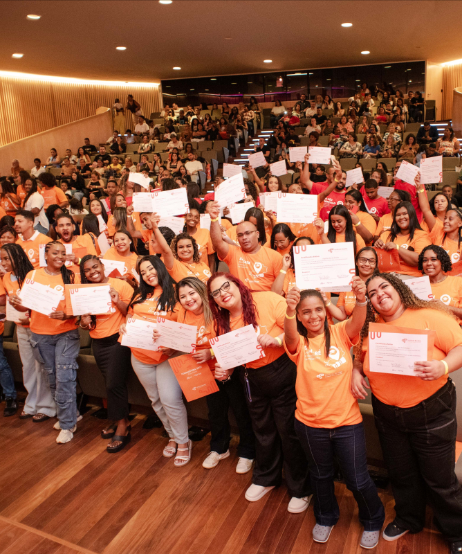 Participantes de uma cerimônia de certificação posam reunidos no auditório, segurando seus certificados com entusiasmo. Os formandos, vestidos com camisetas laranja, ocupam a frente da sala, enquanto o público acompanha a celebração nas arquibancadas ao fundo. A cena transmite sentimento de orgulho coletivo e comemoração.