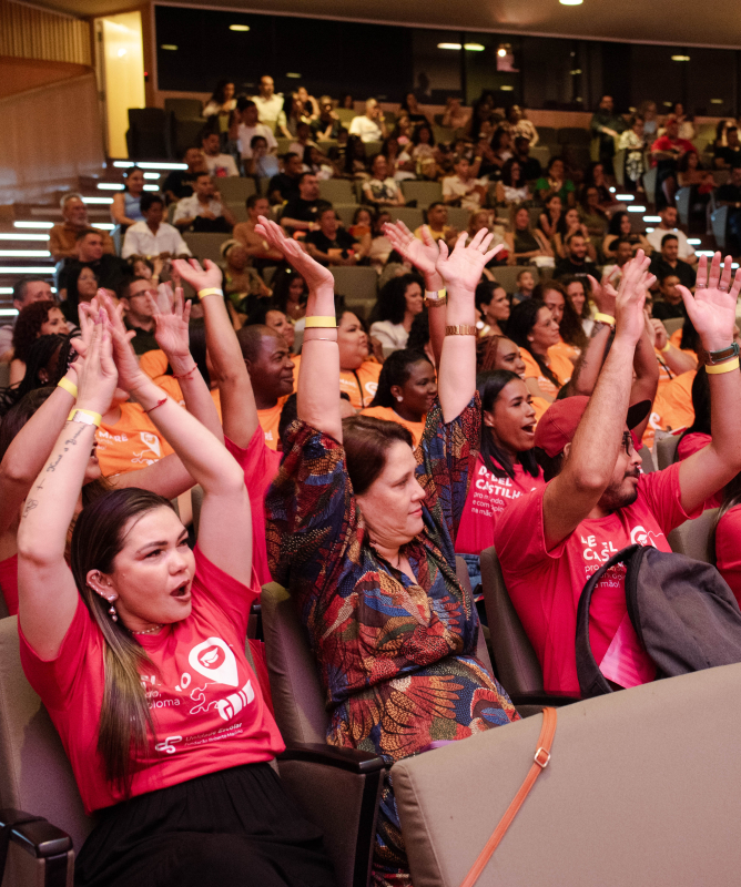 Auditório cheio de pessoas sentadas em poltronas, participando de um evento coletivo. Em primeiro plano, várias pessoas vestem camisetas vermelhas e levantam os braços acima da cabeça, aplaudindo ou comemorando juntas. As expressões são de entusiasmo e envolvimento. Algumas pessoas usam pulseiras e crachás. Ao fundo, o auditório segue lotado, com público em diferentes níveis de assentos, todos voltados para a frente do espaço.