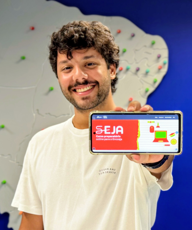Um homem jovem sorri para a câmera enquanto segura um celular com a tela voltada para a frente. Ele usa camiseta branca e está em pé diante de um painel azul. No fundo, há um mapa do Brasil em relevo, com vários pequenos pontos coloridos espalhados pelo território.  Na tela do celular aparece uma página com fundo vermelho e ilustrações coloridas, com o texto em destaque: “SEJA – Curso preparatório online para o Encceja”