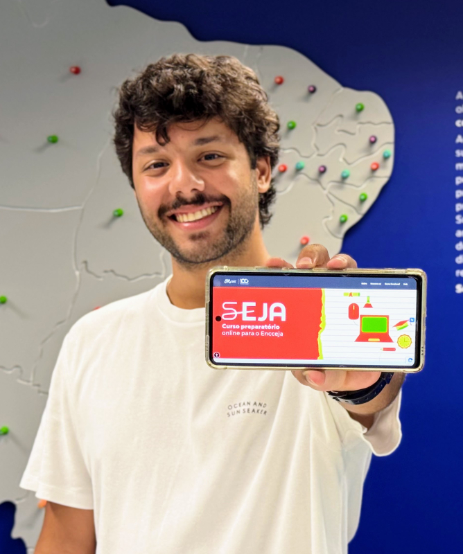 Um homem jovem, sorridente, segura um celular em direção à câmera exibindo a tela do programa SEJA – curso preparatório online para o Encceja. Ele veste camiseta branca e está em frente a um grande mapa do Brasil fixado na parede, marcado com vários pinos coloridos que indicam diferentes localidades.