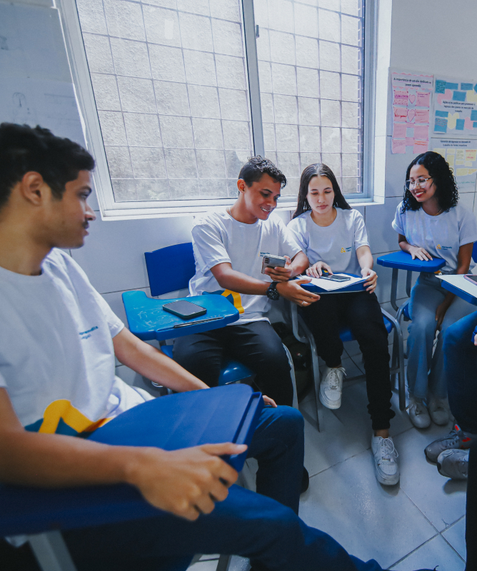 Um grupo de seis jovens está sentado em círculo dentro de uma sala de aula, em cadeiras com apoio para escrita. Todos usam camisetas brancas com um pequeno logotipo colorido. No centro, dois deles compartilham um celular, mostrando algo na tela, enquanto os demais observam com atenção e sorriem. Alguns têm cadernos ou celulares apoiados nas mesas acopladas às cadeiras.