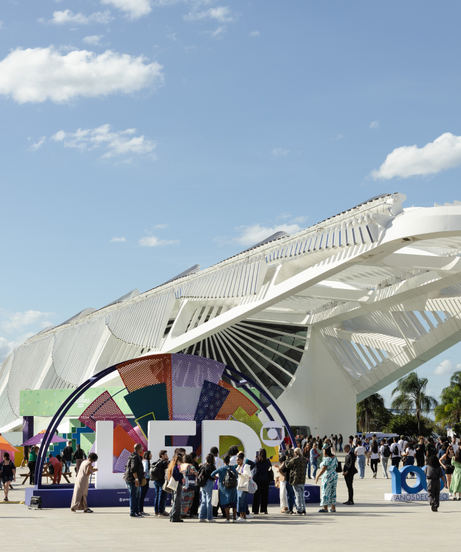 Área externa do Museu do Amanhã, no Rio de Janeiro, com o grande letreiro colorido do Festival LED em primeiro plano. Muitas pessoas circulam pelo espaço em um dia ensolarado, com céu azul e poucas nuvens.