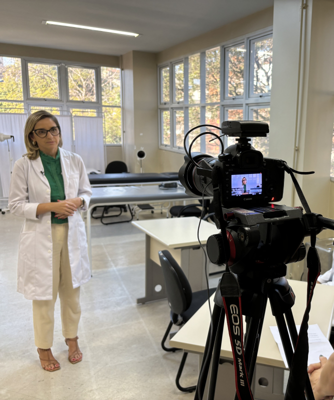 Imagem mostra três pessoas. No primeiro plano, uma jovem opera uma câmera no tripé. O equipamento está direcionado a uma segunda pessoa em pé, que parece estar falando para a câmera. Uma terceira pessoa acompanha a cena sentada ao fundo.