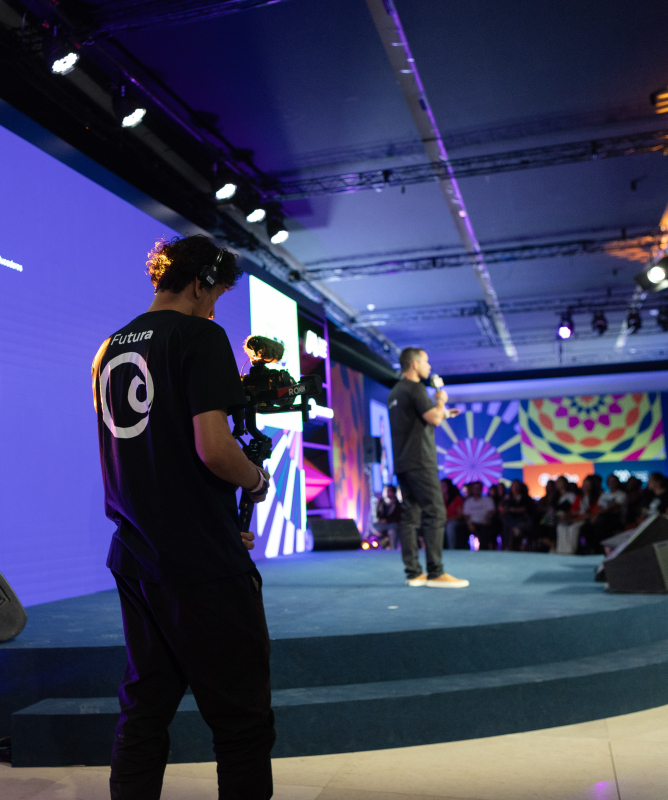 Um cinegrafista filma uma apresentação em um palco durante um evento. Ele aparece em primeiro plano, vestido com camiseta preta e usando equipamentos de gravação presos ao corpo. À frente dele, um homem segura um microfone e fala para uma plateia, embora esteja desfocado ao fundo.  O palco é grande e moderno, iluminado por luzes em tons de azul, roxo e rosa