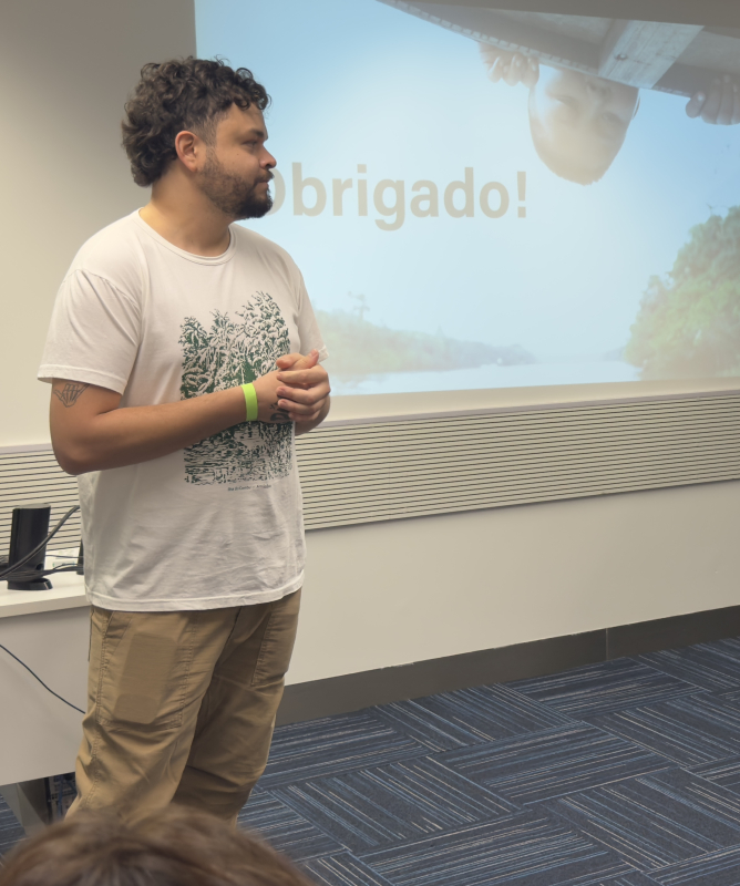 Em uma sala de apresentação, as mesmas duas pessoas aparecem em pé diante de um público. À esquerda, o homem de camiseta branca e calça bege observa a colega enquanto mantém as mãos unidas à frente do corpo. À direita, a mulher de macacão estampado fala gesticulando com as mãos. Atrás deles, um telão projeta a palavra “Obrigado!” sobre uma imagem de rio cercado por vegetação. Ao lado direito há um painel decorativo com fotografia aérea de floresta amazônica. 