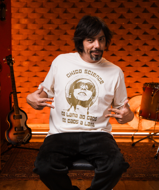 Homem de cabelos médios e escuros posa sentado em um estúdio musical iluminado por luzes alaranjadas. Ele usa camiseta clara estampada com a frase “Chico Science” e aponta para a própria roupa com as duas mãos, em clima descontraído. Ao fundo, aparecem instrumentos musicais, como bateria, guitarra e amplificadores, além de uma parede com tratamento acústico em relevo geométrico. A cena transmite um ambiente criativo e acolhedor, ligado à música e à produção artística.