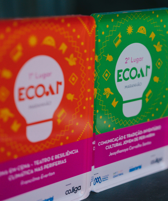 A imagem mostra troféus do prêmio ECOAR Maranhão, organizados lado a lado sobre uma mesa. Em destaque, aparecem premiações de 1º, 2º e 3º lugar, cada uma com cores vibrantes diferentes — vermelho, verde e azul — e o símbolo de uma lâmpada ao centro, associado à ideia de criatividade e inovação.