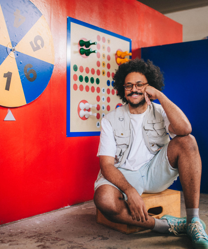 Um homem jovem posa sentado em um pequeno banco de madeira, em um ambiente interno com aparência de estúdio ou espaço expositivo. Ele tem pele morena, cabelo preto volumoso e encaracolado, barba e bigode cheios, além de usar óculos de armação fina. Ao fundo, há um cenário bastante colorido. A parede principal é pintada de vermelho intenso e traz dois painéis de jogos em estilo lúdico.