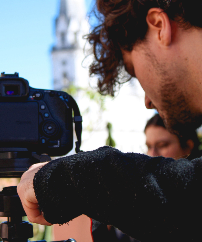 A imagem mostra um jovem operando uma câmera profissional montada em tripé durante uma atividade de gravação ou produção audiovisual. O enquadramento destaca o momento de preparação técnica da filmagem, com foco na câmera e na concentração do operador enquanto ajusta o equipamento.
