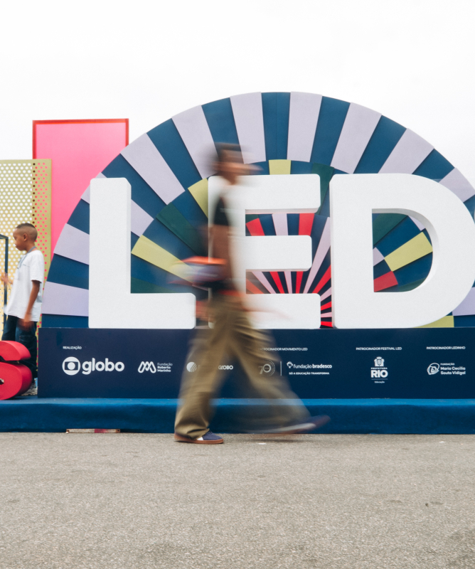 Imagem em ambiente amplo e iluminado, com fundo branco e instalação cenográfica do Festival LED. No centro, há letras gigantes formando a palavra “LED”, em branco, posicionadas diante de um painel semicircular colorido com padrões geométricos em azul, rosa, verde e amarelo. À esquerda, a frase “5 anos” aparece em letras grandes vermelhas sobre um palco azul.