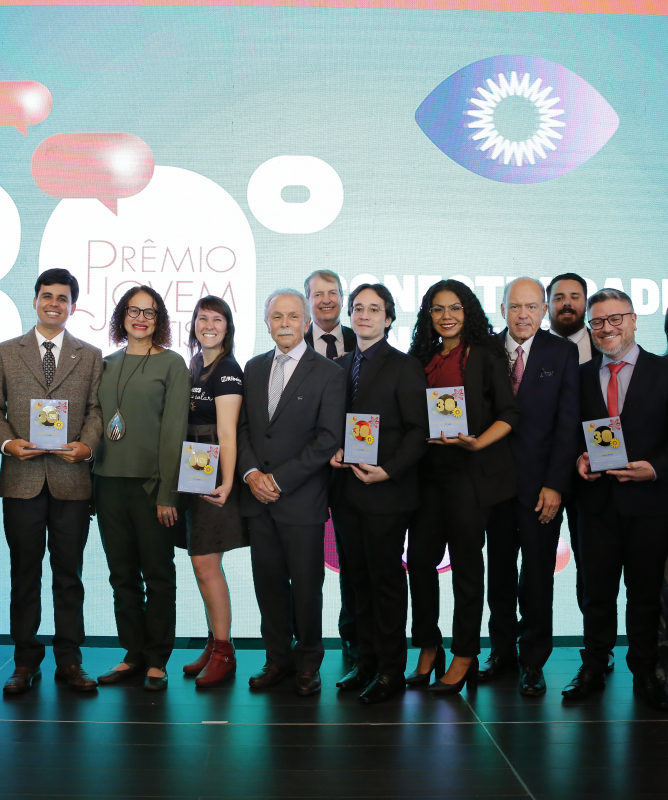 Fotografia de grupo em um palco durante uma cerimônia de premiação. Ao centro, cerca de quinze pessoas, entre jovens estudantes e adultos, posam lado a lado para a foto. Alguns seguram placas ou troféus comemorativos. A maioria está vestida com roupas formais, como ternos, gravatas, vestidos e conjuntos sociais, em cores sóbrias.