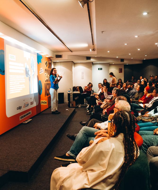 Fotografia de uma palestra em auditório. Em primeiro plano, à esquerda, uma intérprete de Libras aparece em silhueta. No palco, uma mulher com camiseta azul segura um microfone e apresenta conteúdos projetados em uma grande tela. À direita, dezenas de pessoas acompanham atentamente a atividade sentadas em cadeiras organizadas em fileiras. O espaço possui iluminação suave e decoração com a identidade visual do evento em cores vibrantes.