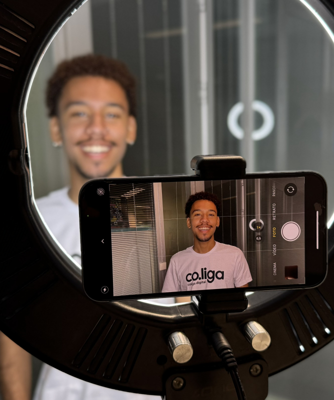 Um jovem aparece sendo fotografado ou gravado em um ambiente interno, diante de um grande ring light. Um celular está posicionado no centro do equipamento e enquadra o jovem, que sorri para a câmera. Na tela do aparelho, é possível ver a imagem dele usando uma camiseta branca com a marca co.liga. Ao fundo, há portas e divisórias de vidro, criando um ambiente com aparência de escritório ou estúdio.