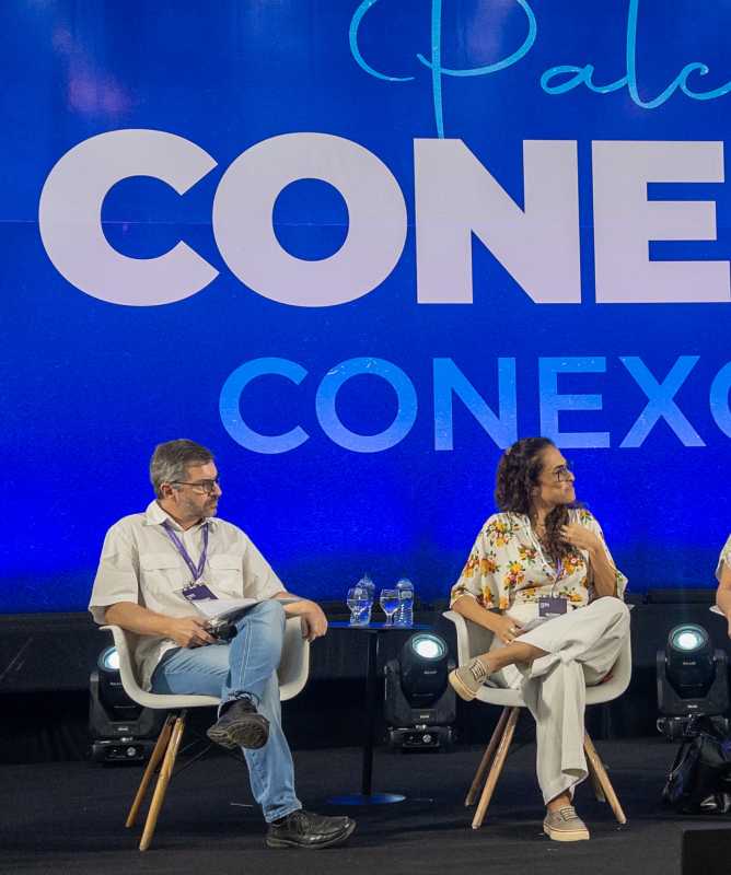 Quatro pessoas sentadas em cadeiras brancas em um palco, com um painel azul ao fundo onde se lê "Palco CONEDU CONEXÕES". Da esquerda para a direita: um homem de camisa bege clara e calça jeans; uma mulher de blusa florida e calça clara; uma mulher de vestido estampado claro e óculos; e um homem de camisa azul, segurando um microfone e falando.