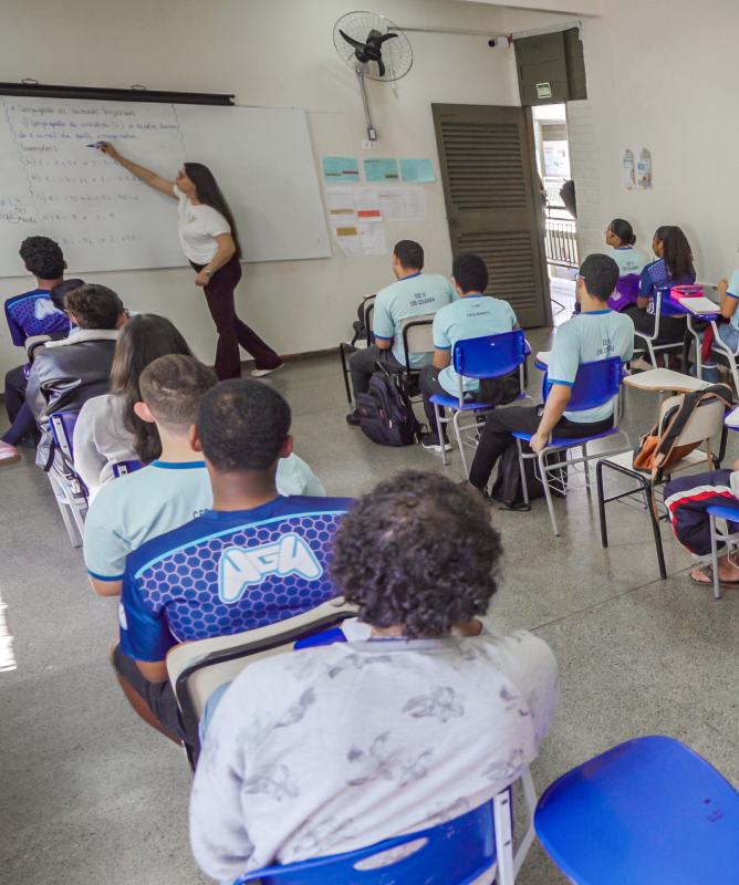 Vista geral de uma sala de aula ampla. À esquerda, uma professora em pé escreve em um quadro branco. Dezenas de estudantes de idades variadas aparecem assentados em carteiras azuis individuais, atentos à explicação, vestindo uniformes escolares em tons de azul e branco ou roupas casuais.