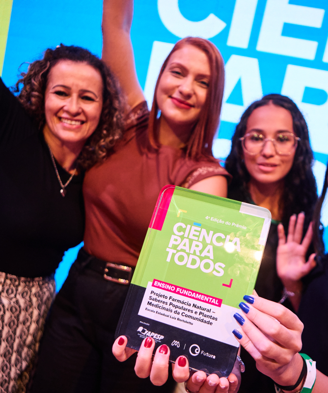 Fotografia colorida em plano médio que registra quatro mulheres sorridentes celebrando em um evento.  No centro do primeiro plano, mãos seguram em destaque uma placa comemorativa retangular. A placa possui design em tons de verde, rosa e preto. Nela, lê-se no topo: “4ª Edição do Prêmio - CIÊNCIA PARA TODOS”.