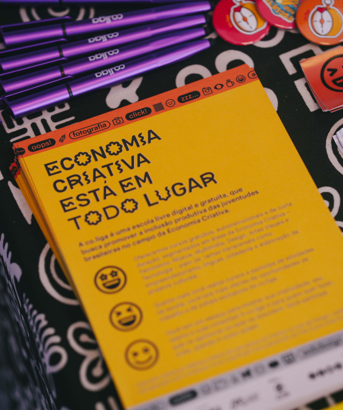 Fotografia colorida em plano detalhe e ângulo superior inclinado (em mergulho), mostrando materiais promocionais e de papelaria organizados sobre uma mesa coberta com um tecido de estampa étnica em preto e branco.