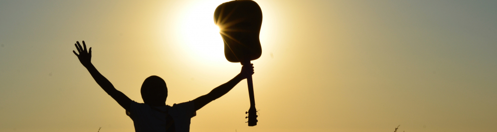 Silhueta de uma pessoa com os braços abertos, segurando um violão com a mão direita de frente para o sol.