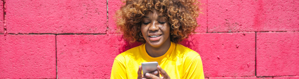 uma jovem de camisa amarela com um celular nas mãos e uma parede de tijolos rosa ao fundo