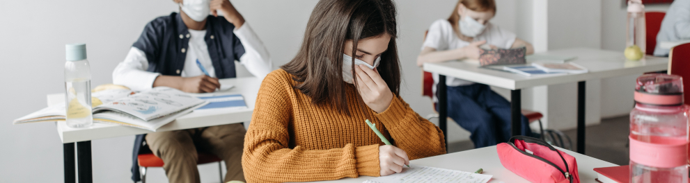 três estudantes usando máscara de proteção sentados nas carteiras escolares de uma sala de aula 
