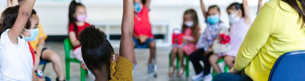 crianças com uma das mãos levantadas sentadas em círculo junto à professora na sala de aula