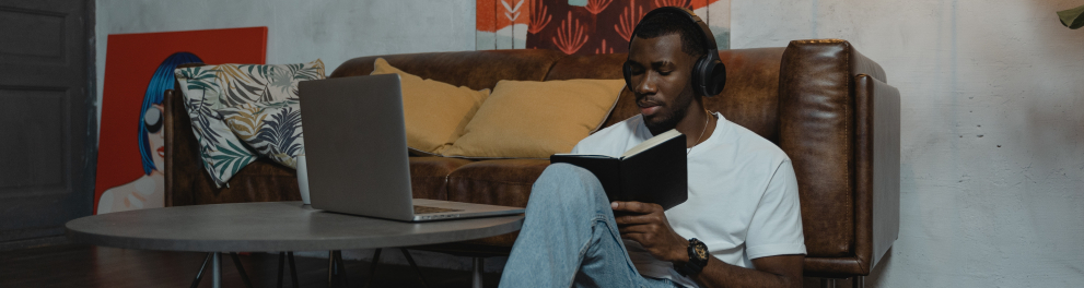 homem negro com fone de ouvido e um livro na mão de frente para um laptop