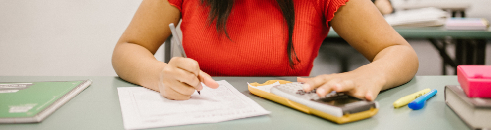 Mulher escreve com um lápis em uma folha de papel e tem uma calculadora ao lado, sobre a mesa.