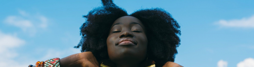 Mulher está de olhos fechados, sorrindo, com as mãos atrás da cabeça. Ao fundo, céu azul.