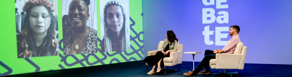 Uma mulher e um homem estão sentados diante de um telão. Nele, três mulheres estão projetadas. Ao fundo da parede, o nome do programa (Debate).