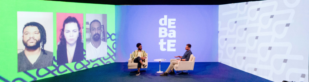 Dois homens estão sentados em poltronas, um de frente para o outro.