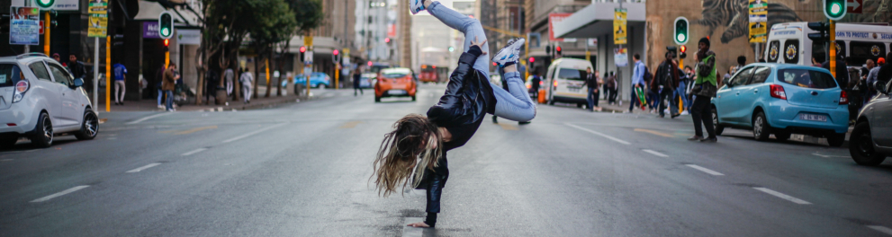 Menina se apoia em uma das mãos no chão em passo de dança, no meio da rua.