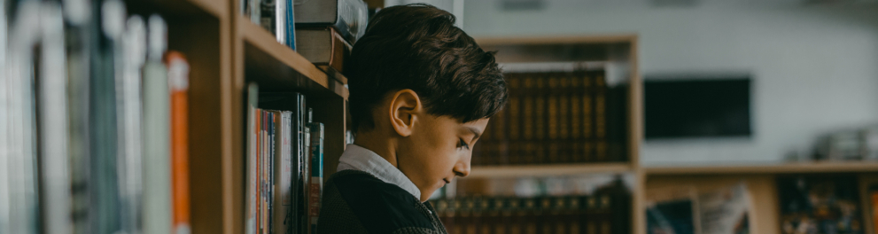 Menino está cabisbaixo, encostado em estante com livros. Parece com medo e/ou triste.