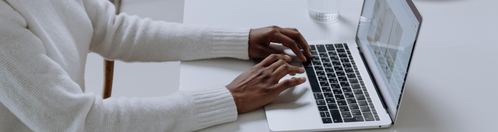 mãos de uma pessoa negra de casaco branco apoiadas na mesa digitando em um laptop