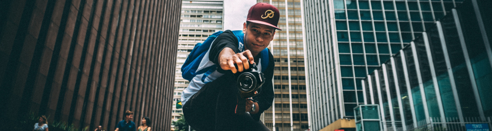 Um homem está agachado no meio da rua, segurando uma câmera fotográfica, dee frente para quem o fotografa.
