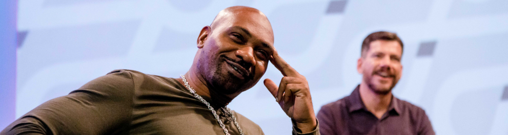 À esquerda, o rapper MV Bill, um homem negro, sorri. À direita, ao fundo, Cristiano Reckziegel, um homem branco, também sorri.