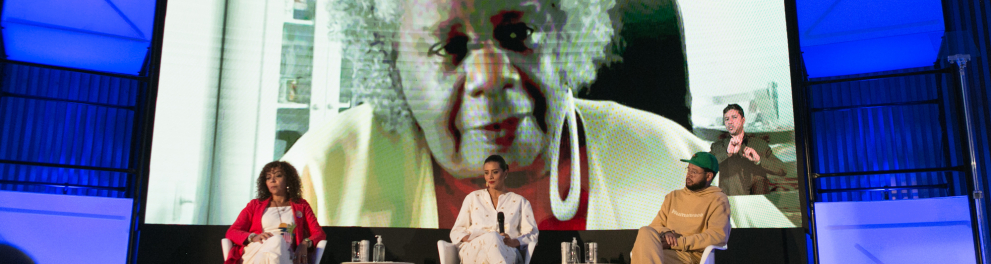 Três pessoas estão sentadas em poltronas. Ao fundo, a imagem de Conceição Evaristo, uma mulher negra, com cabelo crespo e grisalho, é projetada em tamanho maior.