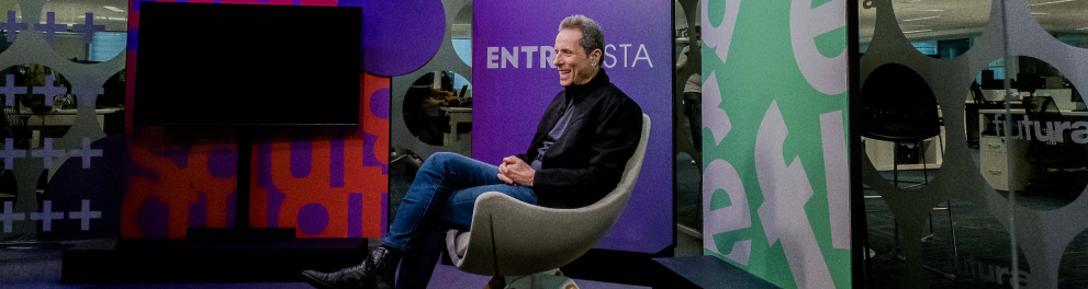 Um homem está sentado em uma poltrona de lado para o fotógrafo que registrou a foto. Ao fundo, a parede conta com paineis nas cores lilás, verde e branca, em que estão o nome do programa "Entrevista".