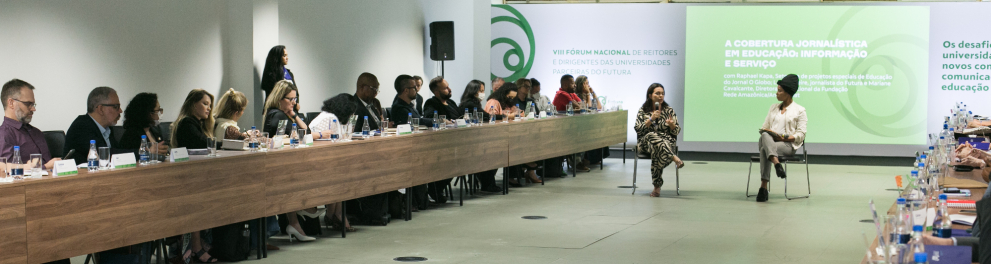 Diversas pessoas estão sentadas à uma longa mesa, no lado esquerdo da imagem. Ao centro, duas mulheres estão sentadas em cadeiras, conversando e sendo escutadas pela plateia. Ao fundo, a logo do Futura.