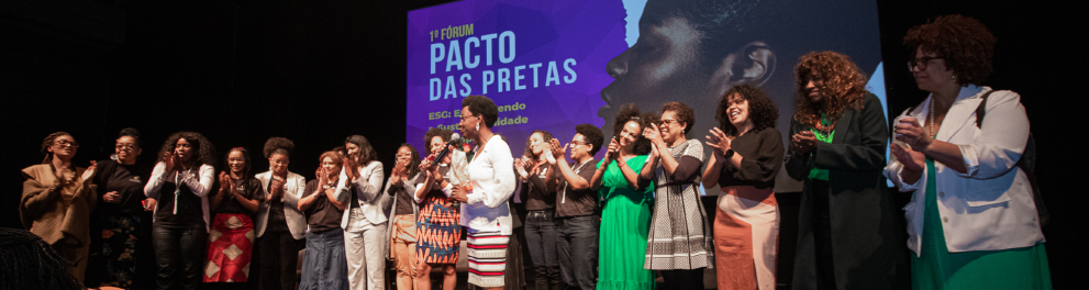 Várias mulheres de pé em um palco aplaudem. Ao fundo, a imagem do Fórum Pacto das Pretas.