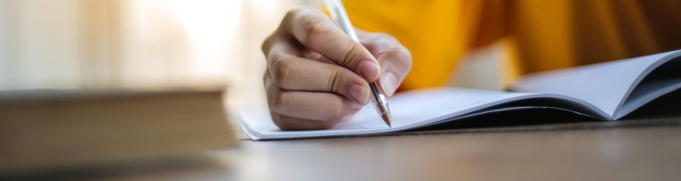 Um homem escrevendo em um caderno com uma caneta. Ele usa um casaco amarelo.