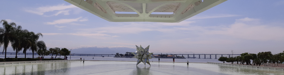 Coqueiros à esquerda, um espelho d'água ao centro, sobre o qual está uma estrela prateada, e, ao fundo, a Baía de Guanabara, com a Ponte Rio-Niterói.