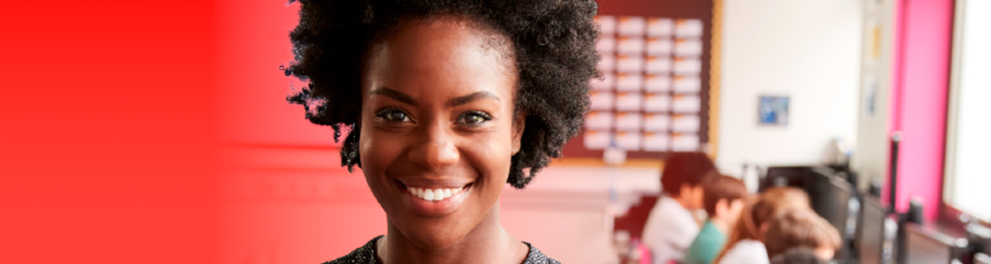 Em primeiro plano, uma mulher negra sorri. Ao fundo, crianças estão sentadas diante de telas de computador.