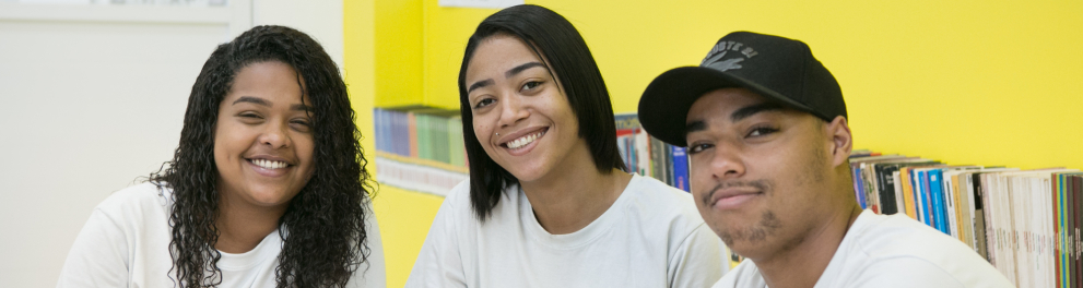 Três jovens sorriem. São duas meninas (à esquerda) e um menino. Todos usam camisas brancas.