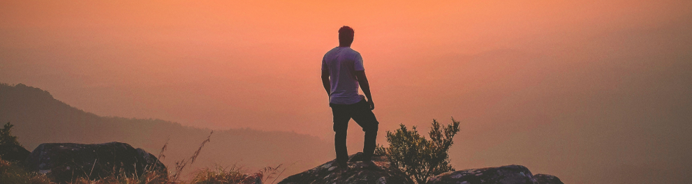 Um rapaz está de pé sobre a uma montanha, contemplando o horizonte em que o sol se põe.
