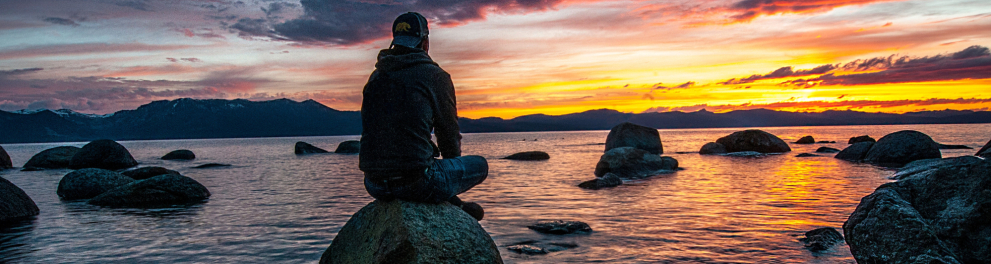 Um homem está sentado sobre uma pedra, de frente para o mar, assistindo ao por do sol.
